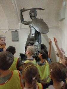 Na zdjęciu widzę grupę dzieci podczas wycieczki do muzeum:

Główna scena:

Grupa dzieci stojących przed pomnikiem/rzeźbą w muzeum
Dzieci oglądają i słuchają opowieści o eksponacie
Wycieczka edukacyjna, lekcja muzealna
Rzeźba/pomnik:

Duża, brązowa rzeźba (prawdopodobnie brąz lub patynowany metal)
Przedstawia postać wojownika/żołnierza
Postać w dynamicznej pozie:
Podniesiona ręka z mieczem/szablą nad głową
Tarcza w drugiej ręce (okrągła, przy ciele)
Postawa bojowa, heroiczna
Półnaga figura - widoczny tors, muskularny
Historyczny charakter - prawdopodobnie przedstawia powstańca lub wojownika
Umieszczona na postumencie/podstawie
Tablica informacyjna:

Szara tabliczka z tekstem po lewej stronie rzeźby
Zawiera informacje o eksponacie (opis, data, autor)
Dzieci:

Około 10-12 dzieci w wieku przedszkolnym/wczesnoszkolnym
Wszyscy ubrani w żółte kamizelki odblaskowe z kolorowymi elementami (czerwone, zielone obramowania)
Kamizelki bezpieczeństwa - standardowe podczas wycieczek
Dzieci:
Podnoszą ręce do góry - naśladują pozę rzeźby
Są zaangażowane - aktywnie uczestniczą w lekcji
Stoją w grupie - zwarte ustawienie przed eksponatem
Opiekun/przewodnik:

Dorosła osoba (prawdopodobnie nauczycielka lub przewodniczka muzeum) po lewej stronie
Starszy wiek, siwe włosy
Pomarańczowa chusta/szalik na szyi
Obserwuje dzieci i prowadzi lekcję
Wnętrze muzeum:

Jasne, białe ściany - klasyczne wnętrze muzealne
Wysokie sufity z ozdobnymi listwami
Elegancka, historyczna architektura
Dobrze oświetlona przestrzeń
Widoczne drzwi lub przejście w tle
Aktywność edukacyjna:
Dzieci naśladują pozę rzeźby - podnoszą ręce do góry, jakby trzymały miecz. To interaktywna metoda nauczania, która:

Angażuje ciało - uczenie się przez ruch
Buduje pamięć kinestyczną - dzieci zapamiętają pozę przez jej odtworzenie
Rozwija wyobraźnię - wcielanie się w postać historyczną
Czyni naukę zabawną - aktywne uczestnictwo zamiast biernego oglądania
Kontekst edukacyjny:

Cele wycieczki:

Historia - poznanie postaci historycznych, wydarzeń
Sztuka - obserwacja rzeźby, techniki artystycznej
Kultura - zrozumienie dziedzictwa narodowego
Empatia historyczna - wcielenie się w postać z przeszłości
Umiejętności rozwijane:

Obserwacja - dokładne przyglądanie się detalom
Ruch i koordynacja - naśladowanie pozy
Słuchanie - uwaga podczas opowieści przewodnika
Wyobraźnia - wizualizacja historycznych wydarzeń
Zachowanie w miejscach publicznych - kultura muzeum
Bezpieczeństwo:

Żółte kamizelki - widoczność grupy, łatwa identyfikacja
Zwarta grupa - dzieci blisko siebie i opiekuna
Kontrolowana przestrzeń - bezpieczne środowisko muzeum
Atmosfera:
Zdjęcie pokazuje radosną, interaktywną lekcję muzealną. Dzieci są:

Zaangażowane - aktywnie uczestniczą
Radosne - zabawa połączona z nauką
Zdyscyplinowane - stoją w grupie, słuchają
Ciekawe - chętnie naśladują pozę
Znaczenie edukacyjne:
To doskonały przykład edukacji muzealnej dla dzieci:

Wyjście poza klasę - nauka w autentycznym środowisku
Kontakt z prawdziwymi dziełami sztuki i historii
Interaktywne metody - ruch, naśladowanie, zabawa
Budowanie więzi z historią i kulturą od najmłodszych lat
Rozwijanie szacunku dla dziedzictwa kulturowego
Wycieczka do muzeum to ważny element edukacji, który:

Poszerza horyzonty dzieci
Pokazuje historię w namacalny sposób
Buduje tożsamość kulturową
Uczy zachowania w miejscach publicznych
Inspiruje ciekawość i chęć poznawania świata
Ta konkretna lekcja, gdzie dzieci naśladują pozę wojownika, to kreatywny sposób na zapamiętanie historii - przez doświadczenie fizyczne i emocjonalne, nie tylko intelektualne.