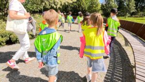 Na zdjęciu widzę grupę dzieci podczas spaceru po parku lub ogrodzie: Główna scena: Grupa dzieci spacerująca ścieżką w parku Opiekunowie prowadzący grupę Słoneczny, letni dzień Wycieczka plenerowa Dzieci: Około 10-15 dzieci w wieku przedszkolnym/wczesnoszkolnym Wszyscy ubrani w jaskrawozielone/limonkowe kamizelki odblaskowe Kamizelki z szarymi pasami odblaskowymi Idą w luźnej grupie, parami lub pojedynczo Dzieci na pierwszym planie: Chłopiec (po lewej): Zielona kamizelka odblaskowa Niebieska koszulka z krótkim rękawem Jasne, krótkie włosy (blond) Szare spodenki jeansowe Idzie spokojnie Dziewczynka (po prawej): Żółta kamizelka odblaskowa z odblaskowymi pasami Różowa bluza z długim rękawem Jasne włosy związane w kucyk Szare spodenki jeansowe Białe skarpetki Idzie obok chłopca Opiekunowie: Dorosła osoba (prawdopodobnie nauczycielka) po lewej stronie Białe ubranie - jasne spodnie i bluzka Różowe buty sportowe Idzie z boku grupy, nadzoruje dzieci Prawdopodobnie więcej opiekunów w grupie (widoczni dalej) Ścieżka i otoczenie: Nawierzchnia: Szeroka, utwardzona ścieżka - kostka brukowa lub płyty Szaro-beżowa nawierzchnia Gładka, równa - bezpieczna dla dzieci Dobrze utrzymana Elementy architektoniczne: Fontanna/kaskada wodna: Nowoczesna fontanna po prawej stronie ścieżki Ciemny, brązowy materiał (prawdopodobnie metal lub kamień) Woda spływająca kaskadowo Dekoracyjny element wodny Współczesny design Drewniana platforma: Drewniane deski tworzące platformę widokową lub ścieżkę Jasne, naturalne drewno Nowoczesna konstrukcja Prawdopodobnie punkt obserwacyjny Zieleń: Drzewa: Wysokie, dorosłe drzewa w tle Gęsta korona - pełne ulistnienie Zielone liście - letnia pora roku Cień i osłona przed słońcem Trawniki: Zadbane, zielone trawniki Świeża, soczysta trawa Dobrze utrzymane tereny zielone Krzewy: Nisko przycięte krzewy po lewej stronie Ozdobne nasadzenia Część kompozycji parkowej Pogoda i światło: Słoneczny dzień - jasne, naturalne światło Bezchmurne niebo lub lekkie zachmurzenie Ciepła pora roku - lato lub późna wiosna Długie cienie - prawdopodobnie przedpołudnie lub popołudnie Doskonałe warunki do spaceru Kontekst wycieczki: Lokalizacja: Prawdopodobnie: Park miejski - nowoczesny, zadbany Ogród botaniczny Teren rekreacyjny Skwer z elementami wodnymi Miejsce dostosowane do spacerów i edukacji Cel spaceru: Edukacyjny: Kontakt z naturą - obserwacja roślin, drzew Nauka o środowisku - ekosystem parku Elementy wodne - fontanny, kaskady Architektura krajobrazu - jak projektuje się parki Rekreacyjny: Ruch na świeżym powietrzu - aktywność fizyczna Relaks - spokojny spacer Integracja grupy - wspólne doświadczenie Odpoczynek po zwiedzaniu muzeum Bezpieczeństwo: Kamizelki odblaskowe: Wysoka widoczność - jaskrawe kolory Łatwa identyfikacja grupy Bezpieczeństwo w ruchu - widoczność dla innych Spójność grupy - wszyscy wyglądają podobnie Opieka: Opiekunowie wśród dzieci i z boku Kontrolowana grupa - dzieci idą razem Bezpieczna trasa - szeroka ścieżka, brak ruchu samochodowego Odpowiednia liczba opiekunów - zapewnienie bezpieczeństwa Organizacja spaceru: Luźna formacja - dzieci nie muszą iść w szeregu Naturalne tempo - każde dziecko w swoim rytmie Swobodna atmosfera - brak sztywnych reguł Komfort - dzieci mogą rozmawiać, obserwować Umiejętności rozwijane: Fizyczne: Chodzenie - wytrzymałość, kondycja Koordynacja - poruszanie się po różnych nawierzchniach Orientacja w przestrzeni - nawigacja w parku Równowaga - chodzenie po ścieżkach Poznawcze: Obserwacja przyrody - rośliny, drzewa, woda Rozpoznawanie elementów - fontanny, ścieżki, architektura Zrozumienie środowiska - jak funkcjonuje park Pamięć - zapamiętywanie trasy, miejsc Społeczne: Chodzenie w grupie - dostosowanie tempa Rozmowy z rówieśnikami - integracja Słuchanie poleceń - posłuszeństwo opiekunom Bezpieczeństwo grupowe - trzymanie się razem Atmosfera: Zdjęcie przedstawia relaksujący, przyjemny spacer: Dzieci spokojne - nie biegają, nie krzyczą Piękna pogoda - zachęca do przebywania na zewnątrz Zadbane otoczenie - estetyczne, bezpieczne Swobodna atmosfera - brak pośpiechu, stresu Znaczenie pedagogiczne: Spacer jako element edukacji: To ważna część programu wycieczki, ponieważ: Równoważy aktywności - po zwiedzaniu muzeum (siedzenie, koncentracja) dzieci potrzebują ruchu Kontakt z naturą - uzupełnienie edukacji kulturalnej o przyrodniczą Relaks - odpoczynek po intensywnych zajęciach Integracja - czas na swobodne rozmowy, budowanie relacji Korzyści dla dzieci: Fizyczne: Ruch - aktywność fizyczna po siedzeniu Świeże powietrze - tlen, witamina D ze słońca Rozciąganie - po statycznych pozycjach w muzeum Energia - rozładowanie napięcia Emocjonalne: Relaks - spokojne, przyjemne otoczenie Radość - przebywanie na zewnątrz Poczucie wolności - przestrzeń, ruch Komfort - przyjemna temperatura, piękne otoczenie Społeczne: Rozmowy - czas na dzielenie się wrażeniami z wycieczki Przyjaźnie - spacer obok kolegów/koleżanek Wspólne doświadczenie - budowanie wspomnień grupowych Integracja - nieformalny czas razem Integracja z całą wycieczką: Struktura dnia: Muzeum - edukacja kulturalna, historia, sztuka (siedzenie, koncentracja) Warsztaty plastyczne - twórczość, ekspresja (siedzenie, precyzja) Spacer po parku - ruch, natura, relaks (aktywność fizyczna, swoboda) To holistyczne podejście do edukacji: Kultura + natura Koncentracja + ruch Nauka + zabawa Struktura + swoboda Nowoczesny park: Otoczenie pokazuje współczesne podejście do przestrzeni publicznej: Estetyka - piękny design, nowoczesna architektura Funkcjonalność - wygodne ścieżki, dostępność Ekologia - zieleń, woda, naturalne materiały Edukacja - przestrzeń do nauki o środowisku Znaczenie dla rozwoju: Taki spacer to nie tylko "przejście z punktu A do punktu B", ale wartościowe doświadczenie edukacyjne: Dzieci uczą się obserwować otoczenie Rozwijają świadomość ekologiczną Budują pozytywne skojarzenia z naturą Uczą się zachowania w przestrzeni publicznej Rozumieją znaczenie ruchu i świeżego powietrza To zdjęcie pokazuje idealną równowagę w programie wycieczki przedszkolnej/szkolnej - po intensywnej edukacji kulturalnej w muzeum, dzieci mają czas na ruch, relaks i kontakt z naturą. Jaskrawe kamizelki zapewniają bezpieczeństwo, a piękna pogoda i zadbane otoczenie sprawiają, że spacer jest przyjemnością. To moment, gdy dzieci mogą przetwarzać to, czego się nauczyły, rozmawiać o swoich wrażeniach i po prostu cieszyć się byciem razem na świeżym powietrzu.