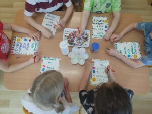 Na zdjęciu widzę grupę dzieci tworzących ręcznie robione kartki z okazji Dnia Mamy i Taty - piękny moment kreatywności i wyrażania miłości:

Główna scena:

Dzieci siedzące wokół stołu - wspólna praca twórcza
Tworzenie kartek okolicznościowych - dla rodziców
Kolorowe dekoracje - mazaki, naklejki, ozdoby
Warsztat plastyczny
Przygotowania do Dnia Mamy i Taty