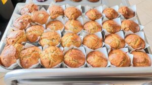 Na zdjęciu widzę efekt końcowy pieczenia - świeżo upieczone muffinki:

Główny element:

Duża metalowa blacha/taca wypełniona świeżo upieczonymi muffinkami
Około 30-35 babeczek ułożonych w rzędach
Wygląd muffinek:

Pięknie wyrośnięte, z charakterystycznymi kopułkowatymi czubkami
Złocistobrązowy kolor - idealnie upieczone
Białe papilotki papierowe
Widoczna delikatna, puszystą tekstura ciasta
Niektóre muffinki mają lekko ciemniejsze plamki - prawdopodobnie z dodatkami (owoce, czekolada lub orzechy)
Równomiernie wyrośnięte, co świadczy o dobrze przygotowanym cieście
Otoczenie:

Białe/jasne kafelki jako powierzchnia
W tle widoczna kolejna forma z muffinkami (brązowe papilotki)
W lewym górnym rogu widoczny fragment żółtego przedmiotu
Profesjonalnie wyglądająca prezentacja
Atmosfera:
Zdjęcie przedstawia satysfakcjonujący rezultat wspólnego pieczenia - dużą ilość pięknie upieczonych, domowych muffinek. Równomierny kolor i kształt świadczą o starannym przygotowaniu. To moment triumfu po całym procesie mieszania, nakładania ciasta i pieczenia - efekt, który z pewnością napawa dumą zarówno dzieci, jak i dorosłych. Widok i zapach świeżo upieczonych babeczek to nagroda za wspólną pracę kulinarną.