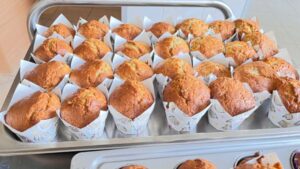 Na zdjęciu widzę pięknie upieczone muffinki prezentowane na tacy:
Główny element:
Duża metalowa blacha/taca z wysokimi krawędziami
Około 24 świeżo upieczonych muffinek ułożonych w równych rzędach (4 rzędy po 6 sztuk)
Wygląd muffinek:
Pięknie wyrośnięte z charakterystycznymi kopułkowatymi czubkami
Złocistobrązowy, karmelowy kolor - idealnie upieczone
Dekoracyjne białe papilotki z wzorem (widoczne małe kolorowe kropki i wzorki - prawdopodobnie misie lub inne urocze motywy)
Papilotki mają charakterystyczny kształt tulipana/korony z wystającymi rogami
Równomierna, puszystą tekstura ciasta
Niektóre muffinki mają delikatne pęknięcia na wierzchu - znak dobrze wyrośniętego ciasta
Widoczne drobne ciemniejsze plamki - prawdopodobnie dodatki (owoce, czekolada lub mak)
Otoczenie:
Jasne, naturalne światło
Białe/jasne kafelki jako tło
W dolnej części zdjęcia widoczna kolejna forma z muffinkami (brązowe papilotki)
Atmosfera:
Zdjęcie przedstawia profesjonalnie wyglądający efekt domowego pieczenia. Dekoracyjne papilotki nadają muffinkom świąteczny, uroczysty charakter - idealne na przyjęcie, urodziny lub specjalną okazję. Równomierny kolor i kształt wszystkich babeczek świadczą o starannym przygotowaniu i doświadczeniu. To moment dumy z osiągniętego rezultatu - piękne, apetyczne wypieki gotowe do podania lub podzielenia się z innymi.