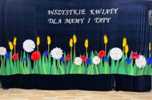 Na zdjęciu widzę pięknie przygotowaną scenę na uroczystość z okazji Dnia Mamy i Taty:

Główna scena:

Scena teatralna - przygotowana na występ dzieci
Dekoracje kwiatowe - ręcznie wykonane
Napis okolicznościowy - dedykacja dla rodziców
Przygotowania do akademii/występu
Sala przedszkolna lub szkolna
Kurtyna:

Charakterystyka:

Granatowa kurtyna - ciemny, głęboki niebieski kolor
Materiał - prawdopodobnie aksamit lub ciężka tkanina
Pełna szerokość sceny - od lewej do prawej
Fałdy - pionowe, równe
Zamknięta - zasłania scenę przed występem
Funkcja:

Tło dla dekoracji - kontrastuje z kolorowymi kwiatami
Zasłona - ukrywa scenę przed występem
Elegancja - nadaje uroczysty charakter
Napis:

Treść:
"WSZYSTKIE KWIATY
DLA MAMY I TATY"

Charakterystyka:

Biały kolor - kontrastuje z granatowym tłem
Duże litery - dobrze widoczne
Czcionka - elegancka, ozdobna (prawdopodobnie serif)
Dwie linie - wyśrodkowane
Umiejscowienie - górna część kurtyny, centralnie
Znaczenie:

Dedykacja - dla rodziców
Nawiązanie do dekoracji - "wszystkie kwiaty" = dekoracje na dole
Okoliczność - Dzień Mamy i Taty
Miłość - wyrażenie uczuć dzieci do rodziców
Dekoracje kwiatowe:

Rozmieszczenie:

Dolna część kurtyny - wzdłuż całej szerokości
Równomiernie rozłożone - harmonijnie
Różne rodzaje kwiatów - różnorodność
Trawa u dołu - tło dla kwiatów
Trawa:

Charakterystyka:

Zielony kolor - jasny, soczyście zielony
Materiał - prawdopodobnie bibuła lub papier
Kształt - pionowe, spiczaste źdźbła
Gęsta - wiele źdźbeł obok siebie
Wzdłuż całej kurtyny - od lewej do prawej