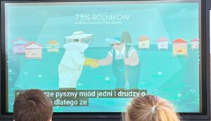 Na zdjęciu widzę kontynuację prezentacji edukacyjnej o pszczołach:

Główny element - ekran/projekcja:

Duży ekran wyświetlający animację edukacyjną
Dominujący miętowo-turkusowy kolor tła
Profesjonalna, minimalistyczna grafika w stylu flat design
Treść na ekranie:

Nagłówek:

"75% ROLNIKÓW" (duży, biały tekst)
"WSPÓŁPRACUJE Z PSZCZEŁAMI" (mniejszy tekst pod spodem)
Ilustracja:

Postać pszczelarza - sylwetka w białym stroju ochronnym z charakterystyczną maską/siatką na głowie
Postać pszczoły - duża, antropomorficzna pszczoła w czarno-żółte paski
Pszczelarz i pszczoła podają sobie ręce/łapki - symbol współpracy
W tle widoczne kolorowe ule (różowe, żółte, białe, pomarańczowe) ustawione w rzędzie
Delikatne białe kwiatki/płatki śniegu unoszące się w powietrzu
Napisy dolne (polskie):

"ze pyszny miód jedni i drudzy o... dlatego że"
Widzowie:

Dwoje dzieci oglądających prezentację
Widoczne z tyłu głowy - brązowe włosy
Jedno dziecko z jasną kokardką we włosach
Atmosfera:
Slajd przedstawia ważną informację edukacyjną o symbiozie między rolnikami a pszczołami. Grafika pokazuje partnerstwo i współpracę (uścisk dłoni) między człowiekiem a naturą. Statystyka "75% rolników współpracuje z pszczołami" podkreśla znaczenie pszczół w rolnictwie i zapylaniu roślin. To część większej prezentacji o znaczeniu pszczół w ekosystemie i gospodarce.