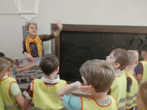 Na zdjęciu widzę przewodniczkę muzealną prowadzącą interaktywną lekcję dla grupy dzieci:

Przewodniczka/edukatorka:

Kobieta stojąca przy eksponacie muzealnym
Żółta kamizelka odblaskowa - taka sama jak dzieci (identyfikacja grupy)
Pomarańczowa chusta/szalik na szyi
Brązowa bluzka z długim rękawem
Jasne włosy związane
Dynamiczna postawa - podniesiona ręka, gestykuluje
Entuzjastyczna mimika - zaangażowana w opowieść
Prowadzi interaktywną prezentację
Dzieci:

Grupa około 8-10 dzieci w wieku przedszkolnym/wczesnoszkolnym
Wszyscy w żółtych kamizelkach odblaskowych z kolorowymi obramowaniami (czerwone, pomarańczowe, zielone)
Dzieci:
Patrzą w górę - skupione na przewodniczce i eksponacie
Podnoszą ręce - chcą odpowiedzieć, są zaangażowane
Stoją blisko - zwarta grupa
Są aktywne - uczestniczą w lekcji
Eksponat muzealny:

Duża, ciemna tablica/płyta z napisami
Prawdopodobnie tablica pamiątkowa lub inskrypcja historyczna
Metalowa lub kamienna powierzchnia z wygrawerowanym tekstem
Tekst w języku polskim (widoczne polskie litery)
Drewniana rama wokół tablicy
Umieszczona na ścianie na wysokości wzroku dorosłych
Gablota/witryna:

Szklana gablota po lewej stronie
Zawiera eksponaty (trudno rozróżnić szczegóły)
Prawdopodobnie artefakty historyczne związane z tematem wystawy
Wnętrze muzeum:

Białe ściany - klasyczne wnętrze muzealne
Jasne oświetlenie - dobrze widoczne eksponaty
Klimatyzacja/wentylacja widoczna na suficie
Przestronna sala wystawowa
Profesjonalne, muzealne otoczenie
Metoda nauczania:

Interaktywność:

Przewodniczka zadaje pytania - dzieci podnoszą ręce
Gestykulacja - pokazywanie, wskazywanie
Entuzjazm - energetyczna prezentacja
Kontakt wzrokowy - przewodniczka patrzy na dzieci
Zaangażowanie dzieci:

Aktywne słuchanie - patrzą w górę
Chęć odpowiedzi - podniesione ręce
Ciekawość - skupienie na temacie
Dyscyplina - stoją w grupie, nie rozpraszają się
Kontekst edukacyjny:

Cele lekcji muzealnej:

Historia - poznanie wydarzeń z przeszłości
Czytanie inskrypcji - nauka odczytywania historycznych tekstów
Pamięć historyczna - zrozumienie znaczenia upamiętniania
Kultura - szacunek dla dziedzictwa
Umiejętności rozwijane:

Słuchanie ze zrozumieniem - uwaga podczas opowieści
Myślenie krytyczne - odpowiadanie na pytania
Obserwacja - przyglądanie się detalom
Pamięć - zapamiętywanie informacji
Zachowanie w grupie - kultura muzeum, cierpliwość
Bezpieczeństwo i organizacja:

Żółte kamizelki - łatwa identyfikacja grupy
Zwarta formacja - dzieci blisko przewodniczki
Kontrolowana przestrzeń - bezpieczne środowisko
Jeden opiekun - jasna struktura grupy
Atmosfera:
Zdjęcie przedstawia żywą, dynamiczną lekcję muzealną:

Przewodniczka pełna pasji - widać zaangażowanie w przekazywanie wiedzy
Dzieci zainteresowane - aktywnie uczestniczą
Interakcja dwustronna - nie tylko słuchanie, ale i odpowiadanie
Radość uczenia się - edukacja przez zabawę i dialog
Znaczenie pedagogiczne:

Edukacja muzealna:
To doskonały przykład nowoczesnej edukacji muzealnej dla dzieci:

Nie tylko oglądanie - aktywne uczestnictwo
Pytania i odpowiedzi - dialog zamiast monologu
Dostosowanie do wieku - język i metody odpowiednie dla małych dzieci
Entuzjazm przewodnika - kluczowy dla zainteresowania dzieci
Korzyści dla dzieci:

Kontakt z autentycznymi obiektami - prawdziwa historia, nie tylko książki
Rozwój ciekawości - chęć poznawania przeszłości
Budowanie tożsamości - zrozumienie własnej kultury i historii
Umiejętności społeczne - słuchanie, czekanie na swoją kolej, podnoszenie ręki
Rola przewodnika:
Przewodniczka na zdjęciu to profesjonalny edukator muzealny, który:

Adaptuje treść - dostosowuje język do wieku dzieci
Angażuje emocjonalnie - używa gestów, mimiki, entuzjazmu
Zachęca do interakcji - zadaje pytania, czeka na odpowiedzi
Tworzy bezpieczną przestrzeń - dzieci czują się komfortowo, chcą uczestniczyć
To zdjęcie pokazuje, jak historia może być żywa i fascynująca dla najmłodszych, gdy jest prezentowana z pasją i w interaktywny sposób. Dzieci nie tylko słuchają, ale są aktywną częścią lekcji, co sprawia, że wiedza lepiej się utrwala i buduje pozytywne skojarzenia z nauką i kulturą.