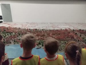 Na zdjęciu widzę grupę dzieci oglądających makietę historycznego miasta w muzeum:

Główna scena:

Troje/czworo dzieci stojących przed dużą makietą
Dzieci patrzą na eksponat z zainteresowaniem
Widok z tyłu - widzimy głowy i plecy dzieci
Dzieci:

Żółte kamizelki odblaskowe z czerwonymi obramowaniami
Dzieci w wieku przedszkolnym/wczesnoszkolnym
Różne fryzury:
Krótkie włosy (chłopcy)
Włosy związane w kucyk (dziewczynka po prawej)
Stoją w rzędzie, blisko siebie
Skupione, spokojne obserwowanie
Makieta miasta:

Rozmiar i konstrukcja:

Bardzo duża, panoramiczna makieta zajmująca całą ścianę
Profesjonalne wykonanie - szczegółowa, realistyczna
Umieszczona na białej podstawie/postumencie
Widok z lotu ptaka na historyczne miasto
Elementy makiety:

Rzeka:

Niebieska wstęga przecinająca miasto
Prawdopodobnie Wisła (biorąc pod uwagę polski kontekst)
Dzieli miasto na dwie części
Mosty łączące brzegi
Zabudowa:

Setki miniaturowych budynków w kolorze terakoty/czerwono-brązowym
Charakterystyczne czerwone dachy - typowe dla historycznych miast europejskich
Różne wysokości budynków - od małych domków po wyższe kamienice
Gęsta zabudowa - historyczne centrum miasta
Wieże kościelne i budynki sakralne - wyróżniające się sylwetki
Zieleń:

Zielone obszary - parki, ogrody, tereny zielone
Drzewa i roślinność wokół miasta
Naturalne otoczenie miasta
Mury obronne:

Widoczne fortyfikacje i mury miejskie
Historyczny charakter miasta-twierdzy
Bastiony i umocnienia
Tło makiety:

Malowane tło przedstawiające krajobraz
Pastelowe kolory - niebo, horyzont
Perspektywa głębi - góry lub wzgórza w oddali
Artystyczne uzupełnienie makiety
Wnętrze muzeum:

Białe ściany - neutralne tło
Jasne oświetlenie - dobrze widoczna makieta
Przestronna sala wystawowa
Profesjonalna ekspozycja
Kontekst historyczny:

Prawdopodobna lokalizacja:
Makieta przedstawia prawdopodobnie historyczne polskie miasto, możliwe opcje:

Warszawa - przed zniszczeniami wojennymi
Kraków - historyczne centrum
Toruń - średniowieczne miasto
Gdańsk - miasto nad rzeką
Inne historyczne miasto z rzeką i fortyfikacjami
Cel edukacyjny:
Makieta pokazuje:

Urbanistykę historyczną - układ ulic, placów, budynków
Architekturę - style budowli, charakterystyczne elementy
Rozwój miasta - jak wyglądało w przeszłości
Geografię - położenie nad rzeką, otoczenie naturalne
Fortyfikacje - system obronny miasta
Wartość dydaktyczna:

Dla dzieci:

Wizualizacja historii - miasto "na żywo", nie tylko na obrazkach
Perspektywa z lotu ptaka - zrozumienie układu przestrzennego
Skala i proporcje - porównanie wielkości budynków
Szczegóły - możliwość dostrzeżenia małych elementów
Wyobraźnia - jak wyglądało życie w dawnym mieście
Umiejętności rozwijane:

Obserwacja - dostrzeganie detali
Myślenie przestrzenne - zrozumienie układu miasta
Wiedza historyczna - jak zmieniały się miasta
Geografia - znaczenie rzek, lokalizacji
Cierpliwość - spokojne oglądanie, analizowanie
Atmosfera:
Zdjęcie przedstawia moment ciszy i skupienia:

Dzieci pochłonięte oglądaniem - nie rozpraszają się
Spokojne obserwowanie - każde dziecko w swoim tempie
Fascynacja - makieta przyciąga uwagę
Edukacja przez doświadczenie - bezpośredni kontakt z eksponatem
Znaczenie pedagogiczne:

Makiety jako narzędzie edukacyjne:

Trójwymiarowość - lepsze zrozumienie niż płaskie mapy
Szczegółowość - możliwość zobaczenia każdego budynku
Kontekst - miasto w otoczeniu naturalnym
Porównanie - jak miasto wyglądało kiedyś vs. dziś
Korzyści dla dzieci:

Konkretność - historia staje się namacalna
Ciekawość - chęć dowiedzenia się więcej
Pamięć wizualna - łatwiejsze zapamiętanie przez obraz
Inspiracja - może zainteresować historią, architekturą, urbanistyką
Rola w wycieczce:
Ta makieta to prawdopodobnie:

Punkt kulminacyjny wycieczki - główny eksponat
Podsumowanie - wizualizacja tego, o czym dzieci słyszały
Punkt odniesienia - dzieci mogą porównać z obecnym wyglądem miasta
Inspiracja do dyskusji - co się zmieniło, co zostało
To zdjęcie pokazuje, jak muzea mogą być fascynujące dla dzieci, gdy oferują interesujące, wizualne eksponaty. Makieta miasta to coś więcej niż suche fakty historyczne - to okno w przeszłość, które pozwala dzieciom zobaczyć, jak wyglądało życie dawniej, i zrozumieć, jak zmieniały się miasta na przestrzeni wieków. Skupienie dzieci na zdjęciu świadczy o tym, że eksponat naprawdę przykuwa ich uwagę i budzi ciekawość.