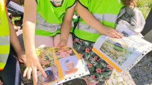Na zdjęciu widzę dzieci oglądające materiały edukacyjne podczas wycieczki: Główna scena: Dwoje lub troje dzieci stojących w kręgu Oglądają książki/albumy edukacyjne Słoneczny dzień na zewnątrz Moment wspólnego czytania i dyskusji Dzieci: Dziecko po lewej: Jaskrawozielona kamizelka odblaskowa z szarymi pasami Jasne, krótkie włosy Ręka wyciągnięta - wskazuje coś w książce Aktywnie uczestniczy w oglądaniu Dziecko w centrum: Zielona kamizelka odblaskowa Jasne spodnie (beżowe lub kremowe) Kolorowa spódnica lub spodenki z kwiatowym wzorem (różowe, czerwone kwiaty na ciemnym tle) Trzyma książkę - pokazuje innym Ręka na książce - wskazuje, pokazuje Dziecko po prawej: Szara bluza z długim rękawem Jasne włosy Stoi z boku, obserwuje Również zaangażowane w oglądanie Książki/materiały edukacyjne: Książka po lewej: Duży album z twardą okładką Żółta/pomarańczowa ramka wokół zdjęć Kolorowe fotografie: Widoczne kwiaty (różowe, czerwone) Rośliny - bujne, zielone Ogród lub park Prawdopodobnie atlas roślin lub przewodnik po ogrodzie Perforacja na krawędzi - może być to materiał z segregatora Profesjonalne, kolorowe zdjęcia Książka po prawej: Podobny format - duży album Żółta/pomarańczowa ramka Fotografie krajobrazowe: Zielone tereny - łąki, pola Drzewa w tle Niebo - jasne, błękitne Prawdopodobnie krajobraz parku lub terenu przyrodniczego Perforacja - materiał edukacyjny z segregatora