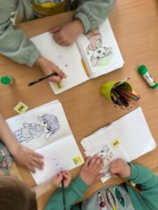 Na zdjęciu widzę scenę przedstawiającą dzieci podczas wspólnych zajęć plastycznych:

Uczestnicy:

Troje dzieci siedzących wokół drewnianego stołu
Dzieci ubrane w miętowe/zielone bluzy
Jedno dziecko ma koszulkę z żółtym motywem (prawdopodobnie postać z kreskówki)
Aktywność:

Dzieci rysują i kolorują w zeszytach/na kartkach
Każde dziecko pracuje nad swoim rysunkiem postaci w stylu anime/manga
Rysunki przedstawiają urocze postacie z dużymi oczami w różnych pozach
Widoczne rysunki:

Góra po prawej: Kolorowana postać w różowej sukience z zielonymi elementami, siedząca w filiżance
Dół po lewej: Szkic dziewczynki z długimi włosami w sukience (niepokolorowany)
Dół po prawej: Postać z rogami/uszkami (częściowo widoczna)
Przybory:

Żółty kubek/pojemnik pełen kolorowych kredek i ołówków
Czarne ołówki do szkicowania
Klej w sztyfcie z zieloną nakrętką
Piórnik z przyborami
Małe żółte naklejki/karteczki na stole
Zeszyty w kratkę
Atmosfera:
Zdjęcie ukazuje typowe dziecięce zajęcia twórcze - spokojną, skoncentrowaną pracę w grupie. Widać, że dzieci są zaangażowane w rysowanie, każde skupione na swoim projekcie. Scena emanuje kreatywnością i współpracą w przyjaznej, edukacyjnej atmosferze.