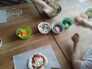 Na zdjęciu widać zaawansowany etap przygotowywania pizzy przez dzieci - moment dodawania składników! Główne elementy: Pizza w trakcie przygotowania (na pierwszym planie): Spód pizzy z sosem pomidorowym Już posypana tartym serem żółtym Leży na papierze do pieczenia z imieniem "Natasza" Bogaty wybór składników w miseczkach: Zielona miska - kolorowa papryka (żółta i czerwona) pokrojona w paski Biała miska - pieczarki pokrojone w plasterki Miętowa miska - salami lub kiełbasa Jasnozielona miska - szpinak lub rukola (świeże liście) Różowa miska - ser mozzarella w kostkach (w tle) Zielona miska - kukurydza (w tle) Metalowa miska - tarty ser żółty (dziecko właśnie bierze z niej ser) Aktywność: Ręce dzieci w ruchu - jedno dziecko sięga po ser z metalowej miski Widoczne ręce kilkorga dzieci wokół stołu Mąka rozsypana na blacie Każde dziecko ma swoje stanowisko Atmosfera: To moment pełnej kreatywności - dzieci mogą wybierać spośród wielu składników i komponować swoje wymarzone pizze. Widać różnorodność opcji, co pozwala każdemu dziecku stworzyć pizzę według własnych preferencji. Profesjonalna organizacja warsztatów z dbałością o szczegóły!