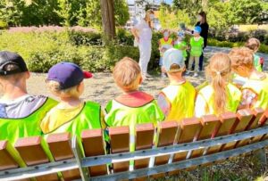 Na zdjęciu widzę grupę dzieci podczas przerwy w parku, słuchających instrukcji od opiekunów: Główna scena: Dzieci siedzą na ławce w parku Dwóch dorosłych opiekunów stoi przed nimi Moment odpoczynku i instruktażu Słoneczny, letni dzień Dzieci na ławce: Liczba i rozmieszczenie: Około 8-10 dzieci siedzących w rzędzie Wiek przedszkolny/wczesnoszkolny Wszyscy w jaskrawożółtych/limonkowych kamizelkach odblaskowych Siedzą spokojnie, zwróceni w stronę opiekunów Widoczne szczegóły: Dziecko skrajnie po lewej: Czarna czapka z daszkiem Jasne włosy wystające spod czapki Patrzy w stronę opiekunów Kolejne dzieci: Fioletowa czapka - dziecko z nakryciem głowy Różowa czapka/bandana - kolorowe akcesoria Jasne, blond włosy - kilkoro dzieci Brązowe/ciemne włosy - różnorodność Kucyki, warkocze - dziewczynki z upięciami Ubrania pod kamizelkami: Bordowa/czerwona koszulka - widoczna u jednego dziecka Żółte, zielone, kolorowe koszulki Letnie, lekkie ubrania Różnorodność kolorów Ławka: Drewniana ławka parkowa z oparciem Jasne, naturalne drewno - sosnowe lub świerkowe Metalowa rama - srebrna/szara konstrukcja Solidna, parkowa ławka Wystarczająco długa dla całej grupy Opiekunowie: Osoba po lewej (kobieta): Białe ubranie - jasne spodnie i bluzka Jasne włosy związane Stoi frontem do dzieci Gestykuluje - prawdopodobnie mówi, instruuje Aktywna postawa - prowadzi rozmowę Osoba po prawej (kobieta): Ciemne spodnie (czarne lub granatowe) Niebieska koszulka z krótkim rękawem Ciemne włosy rozpuszczone Stoi z boku, obserwuje Wspiera pierwszą opiekunkę Dzieci stojące (w tle): Kilkoro dzieci stoi za opiekunami Również w żółtych kamizelkach Część grupy - prawdopodobnie nie zmieściła się na ławce Stoją spokojnie, słuchają Otoczenie - park: Zieleń: Drzewa: Wysokie, dorosłe drzewa w tle Gęste korony - pełne, zielone liście Pień drzewa widoczny po lewej stronie Cień rzucany przez drzewa Dojrzałe drzewostan