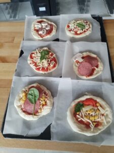 Na zdjęciu widać efekt końcowy pracy dzieci - sześć gotowych do pieczenia pizz ułożonych na blasze! Układ: Dwie blachy piekarnicze (3 pizze na każdej) Każda pizza na osobnym kawałku papieru do pieczenia Widoczne imiona dzieci na papierze (np. "Noel") Różnorodność pizz - każda jest unikalna: Pizza 1 (góra lewa): Sos pomidorowy, pieczarki pokrojone w plasterki Pizza 2 (góra prawa): Sos, tarty ser, świeży szpinak/bazylia Pizza 3 (środek lewa): Sos, ser tarty, liście szpinaku/bazylii Pizza 4 (środek prawa): Sos, ser, salami w plastrach, pieczarki Pizza 5 (dół lewa): Sos, salami, kukurydza, ser, duży liść szpinaku/bazylii jako dekoracja Pizza 6 (dół prawa): Sos, ser tarty, kukurydza, papryka czerwona, liście szpinaku Charakterystyka: Każda pizza odzwierciedla indywidualne preferencje dziecka Różne kombinacje składników Kolorowe i apetyczne kompozycje Widoczna kreatywność w układaniu składników Gotowe do wstawienia do pieca Kontekst: To kulminacja całego procesu warsztatów - od wyrabiania ciasta, przez formowanie spodów, nakładanie sosu, aż po finalne dekorowanie ulubionymi składnikami. Każda pizza to małe dzieło sztuki kulinarnej stworzone przez dziecko!