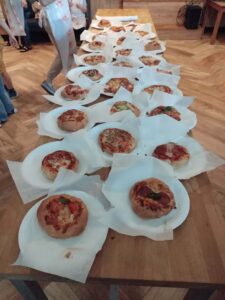 Na zdjęciu widać spektakularną prezentację świeżo upieczonych pizz i wypieków przygotowanych przez dzieci podczas warsztatów!

Układ i prezentacja:

Długi drewniany stół z rzędami białych talerzy i papieru do pieczenia
Pizze i wypieki ułożone w długą linię - prawdziwa wystawa kulinarnych osiągnięć dzieci
Każda pizza/wypiek na osobnym białym talerzu lub papierze
Różnorodność wypieków:

Pizze (większość):

Okrągłe, małe pizze o różnych kompozycjach
Złocisty, rumiany spód
Różne dodatki: salami, pieczarki, papryka, szpinak/bazylia
Ser zapiekany do złocistego koloru
Każda pizza unikalna - odzwierciedla indywidualne preferencje dziecka
Inne wypieki (w tle):

Drożdżówki i bułki w różnych kształtach
Warkocze i sploty z ciasta
Przypominają te z wcześniejszych zdjęć warsztatów
Atmosfera:

Dzieci w tle czekają na swoje wypieki
Widoczne torby z logo piekarni
Drewniane, ciepłe wnętrze
Moment dumy i satysfakcji - prezentacja efektów pracy
Detale:

Widoczne imiona na papierze pod niektórymi wypiekami
Różny stopień zrumienienia - każda pizza wyjątkowa
Apetyczny wygląd wszystkich wypieków
Profesjonalna prezentacja
To kulminacyjny moment warsztatów - wszystkie pizze i wypieki zebrane w jednym miejscu, gotowe do degustacji! Widać ogrom pracy dzieci i różnorodność ich kulinarnej kreatywności. Prawdziwa uczta dla oczu i zapewne także dla podniebienia!