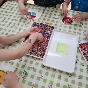 Na zdjęciu widzę fascynujące zajęcia plastyczne - dzieci tworzą obrazki z koralików metodą prasowania (hama beads/perler beads):

Główna scena:

Dzieci przy stole - wspólna praca twórcza
Koraliki do prasowania - tysiące małych, kolorowych koralików
Tworzenie wzorów - układanie koralików na płytkach
Zajęcia plastyczne w przedszkolu lub szkole
Rozwijanie kreatywności i precyzji
Stół:

Obrus:

Wzór - zielono-biała kratka z motywem kwiatowym
Kolor - jasny, świeży
Materiał - prawdopodobnie cerata (łatwa do czyszczenia)
Funkcja - ochrona stołu przed koralkami
Powierzchnia:

Duża - pomieści kilkoro dzieci
Stabilna - bezpieczna dla pracy
Czysta - zadbana
Koraliki (hama beads/perler beads):

Charakterystyka:

Wygląd:

Małe cylindry - około 5 mm średnicy, 5 mm wysokości
Dziurka w środku - charakterystyczna cecha
Plastikowe - z tworzywa sztucznego
Kolorowe - pełna paleta kolorów
Kolory (widoczne):

Czerwony - różne odcienie
Niebieski - jasny, ciemny
Zielony - różne odcienie
Żółty - słoneczny
Pomarańczowy - ciepły
Różowy - jasny, ciemny
Fioletowy - intensywny
Czarny - do konturów
Biały - do wypełnień
Brązowy - naturalny
I wiele innych!
Pojemnik z koralikami:

Charakterystyka:

Duża taca - prostokątna, płaska
Biała - jasna, czysta
Pełna koralików - tysiące sztuk
Pomieszane kolory - tęczowa mieszanka
Centralnie na stole - dostępna dla wszystkich
Funkcja:

Wspólny zasób - dzieci wybierają potrzebne kolory
Łatwy dostęp - każdy może sięgnąć
Organizacja - wszystko w jednym miejscu
Płytki do układania:

Charakterystyka:

Plastikowe płytki - z wypustkami (pinami)
Kształt - kwadratowe, prostokątne
Rozmiar - różne (małe, średnie, duże)
Kolor - przezroczyste lub białe
Wypustki - małe piny, na które nakłada się koraliki
Funkcja:

Szablon - do układania wzorów
Stabilizacja - koraliki trzymają się pinów
Precyzja - równe rzędy i kolumny