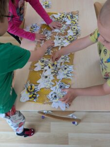 Na zdjęciu widzę grupę dzieci podczas wspólnej zabawy edukacyjnej związanej z pszczołami:

Główna aktywność:

Grupa dzieci (widoczne co najmniej 4 osoby) stojących wokół stołu
Dzieci aktywnie uczestniczą w układaniu/tworzeniu kompozycji
Widok z góry na stół i ręce dzieci
Ubrania dzieci:

Dziecko w różowej/fuksjowej bluzie (lewa strona)
Dziecko w zielonej koszulce (lewa strona, dół)
Dziecko w żółtej koszulce z nadrukiem pszczoły (prawa strona)
Inne dzieci w jasnych ubraniach
Elementy na stole:

Papierowe pszczoły:

Około 30-40 wyciętych papierowych pszczół
Pszczoły są białe z:
Czarnymi paskami na ciele
Żółtymi elementami
Przezroczystymi/białymi skrzydłami
Czarnymi oczkami i uśmiechami
Wszystkie pszczoły są płaskie (2D), wycięte z papieru
Żółte tło:

Żółty papier/karton z wzorem plastra miodu (sześciokąty)
Ułożony w pasie pośrodku stołu
Tworzy "ścieżkę" lub "plaster" dla pszczół
Stół:

Jasny, drewniany blat
Duża powierzchnia robocza
Dodatkowe elementy:

Widoczne nożyczki i inne przybory do pracy plastycznej
Kubek/pojemnik na dole zdjęcia
Atmosfera:
Zdjęcie przedstawia wspólną, kooperacyjną zabawę edukacyjną. Dzieci razem układają pszczoły na plastrze miodu, tworząc dużą, zbiorową kompozycję. To doskonały przykład:

Pracy zespołowej - wszystkie dzieci współpracują przy jednym projekcie
Nauki przez zabawę - manipulowanie elementami, układanie wzorów
Rozwijania motoryki małej - precyzyjne układanie małych elementów
Edukacji przyrodniczej - wizualizacja życia pszczół w ulu
Projekt angażuje wszystkie dzieci jednocześnie, uczy współpracy, dzielenia się przestrzenią i wspólnego tworzenia. Każde dziecko może dodać swoje pszczoły do wspólnego "ula", co daje poczucie przynależności do grupy i wspólnego osiągnięcia. To kulminacyjny moment projektu edukacyjnego o pszczołach - dzieci łączą swoją wiedzę i prace w jedną, dużą instalację.