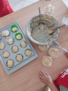 Na zdjęciu widzę scenę przedstawiającą przygotowywanie muffinek lub babeczek:

Główna aktywność:

Przygotowanie ciasta do pieczenia muffinek
Widoczne dzieci w czerwonych koszulkach uczestniczące w procesie
Elementy na stole:

Forma do muffinek (po lewej):

Metalowa forma na 12 babeczek
Większość foremek wypełniona jasnym ciastem
Dwie foremki mają zielone papilotki, pozostałe brązowe/czarne
Ciasto już rozłożone i gotowe do pieczenia
Miska z ciastem (po środku):

Duża metalowa miska z resztkami jasnego ciasta
Dwie łyżki/szpatułki w misce
Ciasto ma gęstą, kremową konsystencję
Dodatkowe elementy:

Trzy białe papilotki/foremki z ciastem leżące obok na stole
Jedna papilotka z ciastem w tle
Drewniany stół jako powierzchnia robocza
Widoczna biała kafelkowa podłoga w tle
Ręka dziecka w dolnej części zdjęcia
Atmosfera:
Zdjęcie dokumentuje proces wypełniania foremek do muffinek ciastem - prawdopodobnie warsztaty kulinarne dla dzieci. Widać, że większość pracy jest już wykonana, a ciasto zostało równomiernie rozdzielone do foremek. Scena emanuje praktyczną nauką gotowania i radością z tworzenia własnych wypieków. To moment tuż przed włożeniem babeczek do piekarnika.