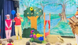 Scena:
Na scenie stoi teatr dla dzieci z kolorowymi kostiumami i dekoracjami.

Aktorzy:
Dwoje dzieci w kostiumach - stojące na scenie
Dziewczynka w różowej i czerwonej koszulce
Chłopiec w żółtej koszulce i czerwonych spodniach
Dekoracje sceniczne:
Duże drzewo - zielone, z żółtym pniem
Niebieskie niebo - jasne, letnie
Zielona trawa i kwiaty - kolorowe (różowe, żółte, niebieskie)
Drewniane elementy - jak płot, kłody drewna
Zwierzęta/postacie - widoczne po prawej stronie sceny
Publiczność:
Dzieci siedzą na podłodze - przed sceną
Patrzą na przedstawienie
Atmosfera:
Wesoła, kolorowa - idealna dla dzieci
Letnia sceneria - natura, słoneczny dzień
Interaktywna zabawa - dzieci uczestniczą w przedstawieniu
Główne wrażenie:
To teatr dla najmłodszych - pełen kolorów, zabawy i radości. Dzieci na scenie grają postaci w naturze, a publiczność obserwuje z zainteresowaniem.