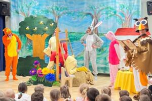 Scena:
Kolorowa scena przedszkolna z bogatymi dekoracjami natury.

Aktorzy na scenie:
Postać po lewej - w pomarańczowo-żółtym kostiumie (ptak/słonecznik?)
Postać w środku - w szarym, puchowym kostiumie (niedźwiedź?)
Postać po prawej - w brązowo-złotym kostiumie z dużymi oczami (sowa?)
Troje dzieci w kostiumach:
Dziewczynka w różowym kostiumie
Chłopiec w żółtym kostiumie
Dziewczynka w czerwonym kostiumie
Dekoracje sceniczne:
Duże zielone drzewo - z brązowym pniem i żółtymi kwiatami
Niebieskie niebo - jasne, letnie
Zielona trawa i łąka - naturalna sceneria
Kolorowe kwiaty - różowe, żółte, niebieskie
Mały domek - po prawej, z czerwonym dachem
Żółty stolik - na scenie
Drewniane elementy - naturalna sceneria
Publiczność:
Dzieci siedzą na podłodze - bardzo blisko sceny
Patrzą z zainteresowaniem - obserwują przedstawienie
Główne wrażenie:
To przedstawienie dla przedszkolaków z trzema zwierzętami w kostiumach (ptak, niedźwiedź, sowa) i dziećmi. Pełne zabawy, kolorów i radości.