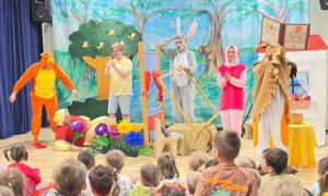 Scena:
Kolorowa scena przedszkolna z bogatymi dekoracjami lasu.

Aktorzy na scenie:
Postać po lewej - w pomarańczowo-żółtym kostiumie (ptak/papuga?)
Postać w żółtym kostiumie - dorosły
Postać w szarym kostiumie - dorosły (niedźwiedź?)
Postać w różowym kostiumie - dziewczynka
Postać w brązowo-złotym kostiumie - dorosły (zwierzę?)
Inne dzieci w kolorowych strojach
Dekoracje sceniczne:
Duże zielone drzewo - z brązowym pniem i żółtymi kwiatami
Niebieskie niebo - jasne, letnie
Zielona trawa i łąka - naturalna sceneria
Kolorowe kwiaty - różowe, żółte, niebieskie
Mały domek - po prawej, z czerwonym dachem
Żółty stolik - na scenie
Drewniane elementy - naturalna sceneria
Publiczność:
Dzieci siedzą na podłodze - bardzo blisko sceny
Patrzą z zainteresowaniem - obserwują przedstawienie
Główne wrażenie:
To przedstawienie dla przedszkolaków z wieloma zwierzętami w kostiumach i dorosłymi aktorami. Pełne zabawy, kolorów i radości. Wszyscy razem bawią się na leśnej łące. 