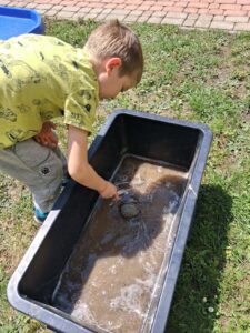 Na zdjęciu chłopiec pochyla się nad czarną plastikową wanną z wodą i błotem, mieszając w niej patykiem. W tle widać trawę i bruk.