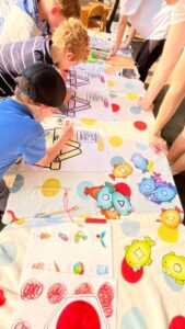 Dzieci malują na długim plakacie 🎨🖍️
Stół - długi, biały:
Widok z góry, wzdłuż stołu

Dzieci (około 4-5 widocznych):
Po obu stronach stołu - pochyleni nad pracą
Ubrania - kolorowe (niebieskie, białe koszulki)
Włosy - różne (jasne, ciemne, kręcone)
Aktywność - malują, rysują
Plakat/papier - biały, długi:
Rozłożony wzdłuż całego stołu

Rysunki dzieci:
Kolorowe kształty:

Żółte koła - słońca, planety
Czerwone kształty - serca, koła
Niebieskie elementy - chmury, kształty
Zielone - różne formy
Postacie/ilustracje:

Kolorowe roboty/potwory - narysowane i wycięte:
Niebieski robot - z antenkami
Czerwony potwór - z zębami
Żółty robot - geometryczny
Zielony potwór - na dole
Styl - dziecięcy, wesoły, kreskówkowy
Wzory:

Czerwone kreski - w kratkę (dół plakatu)
Linie - różne kolory
Abstrakcyjne kształty
Materiały na stole:
Farby/stemple:
Czerwone - okrągłe odciski
Żółte - okrągłe odciski
Niebieskie - widoczne
Zielone - kolorowe elementy
Przybory:
Pędzle - leżą na stole
Kredki/mazaki - kolorowe
Gąbki - do stemplowania
Pojemniki - z farbami
Dorośli:
Kilka osób - w tle, nadzorują
Ubrania - białe, fioletowe koszulki
Główne wrażenie:
To wspólny projekt artystyczny! Dzieci tworzą wielki, kolorowy plakat pełen robotów, potworów i abstrakcyjnych kształtów. Każde dziecko dodaje swoje kolorowe elementy - maluje, stempluje, rysuje. Kreatywna zabawa zespołowa rozwijająca wyobraźnię i współpracę! 