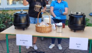 Stoisko z zupami 🍲🥣
Stół:
Blat - jasny, beżowy
Nogi - zielone, metalowe
Rozmiar - długi, prostokątny
Garnki z zupą (2 sztuki):
Lewy garnek:
Kolor - czarny, błyszczący
Typ - podgrzewacz elektryczny do zupy
Pokrywka - czarna, z uchwytem na górze
Napis - "Grochówka 8zł" (zupa grochowa za 8 złotych)
Prawy garnek:
Kolor - czarny, identyczny jak lewy
Typ - podgrzewacz elektryczny
Pokrywka - czarna
Napis - "Żurek 8zł" (żurek za 8 złotych)
Przedmioty na stole:
Środek:
Koszyk wiklinowy - jasny, beżowy
Zawartość - chleb, pokrojony w kromki, biały obrus
Pojemnik metalowy - srebrny, cylindryczny (termos lub pojemnik)
Tabliczki z cenami:
Białe kartki - przy każdym garnku
Napisy - nazwy zup i ceny
Ludzie (dwoje):
Osoba po lewej:
Ubranie - czarna bluza z napisem, niebieskie dżinsy
Buty - białe sportowe
Pozycja - stoi za stołem
Osoba po prawej:
Ubranie - niebieska koszulka polo, niebieskie dżinsy
Buty - białe sportowe
Pozycja - stoi za stołem, trzyma coś w rękach
Otoczenie:
Podłoże:
Kostka brukowa - szara
Tło:
Ściana - beżowa/kremowa
Rośliny - zielone, w doniczkach po bokach
Okno - po lewej stronie
Główne wrażenie:
To stoisko z tradycyjnymi polskimi zupami! Oferuje grochówkę i żurek po 8 złotych za porcję. Dwa duże podgrzewacze utrzymują zupy gorące. W wiklinowym koszyku leży świeży chleb do zup. Dwoje wolontariuszy obsługuje głodnych gości festynu! 