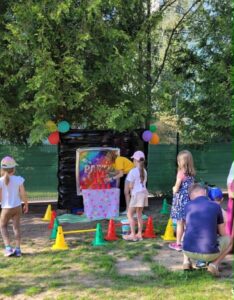 Gra "Rzutki" na festynie 🎯🎈
Główna atrakcja - tablica do rzutek:
Konstrukcja:
Rama - czarna, drewniana, duża
Tablica - kolorowa, z napisem "PIN THE TAIL" (przypnij ogon)
Tło - tęczowe kolory (różowy, żółty, niebieski)
Cel - różowa tablica z kropkami (miejsca do celowania)
Dekoracje:
Balony - kolorowe (zielone, pomarańczowe, różowe) przyczepione do ramy
Paciorki - czarne, wiszące po bokach ramy
Dzieci (około 6-7):
Przed tablicą:
Chłopiec w żółtej koszulce - stoi najbliżej, celuje
Dziewczynka w białej koszulce - obok, z różową czapką
Dziewczynka w białej sukience - po lewej
Dziewczynka w granatowej sukience - po prawej, z wzorami
Dziewczynka z jasnym włosami - stoi z boku
Ubrania:
Kolorowe - żółte, białe, różowe, granatowe
Letnie - sukienki, koszulki, szorty
Czapki - niektóre dzieci mają różowe czapeczki
Przeszkody - pachołki:
Żółte - 3 sztuki (po lewej)
Pomarańczowe - 3 sztuki (po prawej)
Układ - wyznaczają linię startu
Dorośli:
Osoba w granatowej koszulce - klęczy po prawej, pomaga dzieciom
Osoba w różowym - stoi z boku
Otoczenie:
Przyroda:
Drzewa - duże, zielone, gęste liście
Trawa - zielona, świeża
Cień - od drzew
Tło:
Płot/siatka - zielona, za tablicą
Niebo - błękitne, widoczne przez liście
Pogoda:
Słonecznie - jasne światło
Ciepło - letni dzień
Główne wrażenie:
To gra zręcznościowa "Przypnij ogon" (Pin the Tail)! Dzieci rzucają rzutkami w kolorową tablicę, celując w różowe kropki. Żółte i pomarańczowe pachołki wyznaczają linię startu. Kolorowe balony i tęczowa tablica tworzą wesołą atmosferę! Zabawa rozwijająca celność i koordynację! 