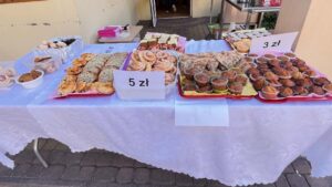Stoisko z ciastami na festynie 🧁🍰
Stół:
Obrus - biały, długi, papierowy
Kostka brukowa - szara, pod stołem
Wypieki od lewej do prawej:
Lewa strona:
Ciastka - małe, różne rodzaje
Brak ceny - widocznej
Środek-lewo:
Drożdżówki - duże, płaskie
Posypka - biała (cukier puder) i brązowa (cynamon)
Cena - "5 zł" (biała kartka)
Środek:
Ślimaczki drożdżowe - zwinięte w spirale
Kolor - złotobrązowe
Nadzienie - widoczne warstwy (może cynamon, mak)
Środek-prawo:
Babeczki/muffiny - w papilotach
Kolor - ciemnobrązowe (czekoladowe)
Polewa - czekoladowa, błyszcząca
Cena - "3 zł" (biała kartka)
Prawa strona:
Babeczki - w kolorowych papilotach
Polewa - różowa, brązowa
Dekoracje - posypki
Tło:
Budynek - beżowa ściana
Drzwi - ciemne, w tle
Inne stoiska - widoczne dalej
Pojemniki - różowe, plastikowe (na górze stołu)
Główne wrażenie:
To stoisko ze świeżymi wypiekami na festynie! Domowe ciasta w atrakcyjnych cenach: drożdżówki po 5 zł, babeczki po 3 zł. Wszystko wygląda świeżo upieczone i apetycznie. Pięknie ułożone na tackach! 