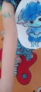 Zdjęcie przedstawia dziecko trzymające rysunek niebieskiego potwora - widok z góry na dziecko z kolorowym tatuażem na ramieniu i dużą czerwoną cyfrą "6" na podłodze.

Dziecko (centrum):

Ramię (lewo, góra):

Jasna skóra
Kolorowy tatuaż tymczasowy (zmywalny) na ramieniu
Kształt czaszki lub cukrowej czaszki (sugar skull)
Kolorowe wzory - pomarańczowy, niebieski, zielony
Dekoracyjne elementy - kropki, wzory
Styl meksykański - Día de los Muertos (Dzień Zmarłych)
Małe ramię dziecka - około 5-8 lat
Ubranie:

Niebieska sukienka lub koszulka
Wzór kwiatowy lub abstrakcyjny
Ciemnoniebieski z jaśniejszymi elementami
Krótki rękaw
Nogi (dół):

Jasne nogi dziecka
Skarpetki w paski (czarno-białe lub czarno-szare)
Poziome paski - klasyczny wzór
Długie skarpetki - do kolan lub wyżej
Stoi na podłodze
Rysunek (centrum, prawo):

Postać na rysunku:

Niebieski potwór - przyjazny, kreskówkowy
Duża głowa z puchatymi, niebieskimi włosami
Intensywny niebieski kolor
Rozczochrane włosy - wystające na boki
Gęste, pusziste - jak futro
Duże, okrągłe oczy
Białe gałki oczne
Czarne źrenice - patrzą w bok
Szeroko otwarte - zdziwiony lub radosny wyraz
Duży, szeroki uśmiech
Białe zęby - widoczne
Szeroki, przyjazny - radosny wyraz
Czarna linia ust
Mały nosek - prosty kształt
Niebieskie ciało
Krótkie ręce - przy ciele
Krótkie nogi - grube, stabilne
Okrągły brzuch - puchaty kształt
Stoi - frontalnie
Styl rysunku:

Kreskówkowy - przyjazny, dziecięcy
Kolorowy - intensywny niebieski
Prosty kontur - czarne linie
Wyraźne kolory - dobrze pokolorowane
Biały papier - okrągły lub owalny kształt
Cyfra "6" (dół, centrum):

Duża czerwona cyfra:

Intensywny czerwony kolor
Duża, gruba cyfra - około 20-30 cm wysokości
Wycięta z papieru lub kartonu
Płaska - leży na podłodze
Czerwony karton - matowy
Cyfra "6" - wyraźna, czytelna
Czarny kontur lub cień wokół cyfry
Ciemny obrys - dodaje głębię
Może naklejka lub malowana
Podłoga (tło):

Powierzchnia:

Jasne drewno - naturalne deski lub panele
Beżowy/kremowy kolor
Gładka powierzchnia - parkiet lub panele
Czysta - zadbana podłoga
Charakterystyka:

Aktywność: Prezentacja rysunku - dziecko pokazuje swoją pracę

Możliwy kontekst:

1. Urodziny:

Cyfra "6" - dziecko ma 6 lat
Urodzinowa dekoracja - czerwona cyfra
Tatuaż tymczasowy - urodzinowa zabawa
Rysunek - urodzinowa aktywność
2. Warsztaty artystyczne:

Rysunek - efekt warsztatów plastycznych
Tatuaż - część zabawy/warsztatów
Cyfra - numer uczestnika lub stacji
Prezentacja pracy - pokazanie efektów
3. Zabawa tematyczna:

Tatuaż meksykański - Día de los Muertos
Niebieski potwór - postać z bajki (może z "Gdzie mieszkają dzikie stwory"?)
Cyfra - element gry lub zabawy
Kostium - pasiaste skarpetki jako część stroju
Postać na rysunku - możliwe inspiracje:

1. "Gdzie mieszkają dzikie stwory" (Where the Wild Things Are):

Niebieski potwór - podobny styl
Puchate włosy - charakterystyczne
Przyjazny wyraz - nie straszny
2. Oryginalna kreacja dziecka:

Własna wyobraźnia - unikalna postać
Niebieski kolor - ulubiony kolor?
Radosny potwór - przyjazny, nie straszny
Tatuaż - znaczenie:

Cukrowa czaszka (sugar skull):

Meksykańska tradycja - Día de los Muertos
Kolorowa, radosna - nie straszna
Edukacja kulturowa - nauka o innych kulturach
Sztuka ludowa - piękne wzory
Tatuaż tymczasowy:

Zmywalny - bezpieczny dla dzieci
Zabawa - dzieci uwielbiają tatuaże
Kreatywność - wyrażanie siebie
Tymczasowy - zmyje się po kilku dniach
Cyfra "6" - możliwe znaczenia:

1. Wiek dziecka:

6 urodziny - dziecko ma 6 lat
Urodzinowa dekoracja - część dekoracji
Sesja zdjęciowa - pamiątkowe zdjęcie
2. Numer uczestnika:

Warsztaty - dziecko nr 6
Zawody - numer startowy
Gra - numer gracza
3. Element edukacyjny:

Nauka liczb - rozpoznawanie cyfr
Matematyka - liczenie, cyfry
Aktywność edukacyjna - nauka przez zabawę
Pasiaste skarpetki - styl:

Klasyczny wzór - czarno-białe paski
Zabawny element - dziecięcy styl
Może część kostiumu - strój tematyczny
Modny akcent - dzieci lubią pasiaste skarpetki
Wartości edukacyjne:

Rozwój artystyczny:

Rysowanie - tworzenie postaci
Kolorowanie - precyzyjne wypełnianie
Kreatywność - wymyślanie postaci
Prezentacja - pokazywanie swojej pracy
Rozwój kulturowy:

Tatuaż meksykański - nauka o innych kulturach
Sztuka ludowa - piękne wzory i tradycje
Otwartość - poznawanie różnorodności
Szacunek - dla innych kultur
Rozwój społeczny:

Prezentacja pracy - dzielenie się efektami
Duma - "Zobacz, co stworzyłem!"
Pewność siebie - pokazywanie swoich umiejętności
Radość tworzenia - satysfakcja z pracy
Aspekt emocjonalny:

Radość - dziecko dumne ze swojej pracy
Kreatywność - wyrażanie siebie przez sztukę
Zabawa - tatuaż, rysunek, cyfra - wszystko jest zabawą
Satysfakcja - "Zrobiłem to!"
Możliwy scenariusz:

Urodziny 6-latka:

Rano: Tatuaż tymczasowy jako prezent/zabawa
Warsztaty artystyczne: Rysowanie potworów
Prezentacja: Dziecko pokazuje swój rysunek
Sesja zdjęciowa: Zdjęcie z rysunkiem i cyfrą "6"
Pamiątka: Piękne wspomnienie z urodzin
Przekaz:

Sztuka jest dla wszystkich - każdy może tworzyć
Kreatywność jest ważna - wyrażaj siebie
Różnorodność kulturowa - poznawaj inne kultury
Radość tworzenia - sztuka to zabawa
Duma z efektów - pokazuj swoje prace
Kolorowe, radosne zdjęcie przedstawiające dziecięcą kreatywność i radość tworzenia! Niebieski potwór jest przyjazny i uśmiechnięty - pokazuje, że:

Potwory nie muszą być straszne - mogą być przyjaciółmi
Kreatywność nie ma granic - wymyślaj własne postaci
Sztuka to zabawa - rysowanie jest fajne
Możesz być dumny ze swojej pracy
Kolorowy tatuaż i pasiaste skarpetki dodają zabawnego, dziecięcego charakteru, a czerwona cyfra "6" może oznaczać specjalny dzień - urodziny lub ważne wydarzenie.

Piękny przykład dziecięcej kreatywności, radości i dumy z własnych osiągnięć artystycznych