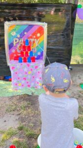 Dziecko bawi się w "Zbij puszkę" 🎯🎉
Dziecko:
Widok - od tyłu
Ubranie - szara bluza
Włosy - jasne, krótkie
Pozycja - stoi, celuje do gry
Ręka - wyciągnięta, trzyma piłkę
Gra "Zbij puszkę":
Tablica/Cel:
Rama - biała, drewniana
Tło - kolorowe, tęczowe (różowe, niebieskie, żółte, fioletowe)
Napis - "PARTY" (duże, białe litery z gwiazdkami)
Puszki - kolorowe (czerwone, niebieskie, żółte) ułożone piramidą
Dół tablicy - różowe konfetti/kropki
Podstawa:
Kolor - biała
Materiał - drewno lub plastik
Stabilna - stoi na ziemi
Otoczenie:
Tło:
Płot - ciemny, drewniany (brązowy/czarny)
Siatka - zielona, za płotem
Balony - kolorowe (fioletowe, tęczowe) po prawej stronie
Podłoże:
Kostka brukowa - szara
Trawa - zielona, po bokach
Mata - kolorowa, pod grą
Główne wrażenie:
To gra zręcznościowa "Zbij puszkę"! Dziecko celuje piłką w kolorowe puszki ułożone na tablicy. Tęczowe tło z napisem "PARTY" dodaje świątecznej atmosfery. Klasyczna zabawa na festynie rozwijająca celność i koordynację!