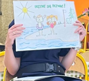 Dziecięcy rysunek przedstawiający scenę nad wodą. U góry widnieje napis "Uczmy się pływać!" w kolorze turkusowym. Na rysunku znajdują się dwie postaci dzieci stojące na pomoście lub brzegu - jedno z krótszymi włosami w fioletowych spodenkach, drugie z dłuższymi, rudymi włosami w różowym stroju kąpielowym. Obie postaci mają uniesione ręce w geście radości. W lewym górnym rogu narysowane jest słońce z promieniami. U dołu widać niebieskie fale symbolizujące wodę. Po prawej stronie widać fragment zielonej postaci, prawdopodobnie kolejnej osoby. Rysunek jest trzymany przez osobę siedzącą na żółtym krześle.