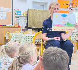 Kobieta w granatowym mundurze straży (z napisem "STRAŻ") siedzi na żółtym krześle i pokazuje dziecięcy rysunek grupie dzieci siedzących przed nią na podłodze. Widoczne są głowy czworga dzieci z blond i ciemniejszymi włosami, które patrzą na prezentowany rysunek.

Rysunek przedstawia postać dziecka stojącą nad wodą, z żółtym słońcem i niebieską chmurą na niebie. U dołu rysunku widnieje napis w kolorze czerwonym. Scena rozgrywa się w jasnym, kolorowym pomieszczeniu przedszkolnym lub szkolnym, z żółtymi krzesłami, białym płotkiem dekoracyjnym w tle oraz tablicami informacyjnymi na ścianach. To wygląda na zajęcia edukacyjne dotyczące bezpieczeństwa nad wodą prowadzone dla dzieci.