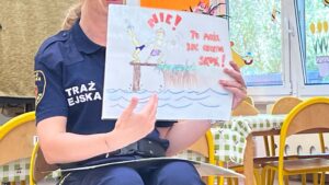 Kobieta w granatowym mundurze z napisem "STRAŻ MIEJSKA" siedzi na krześle i trzyma dziecięcy rysunek. Rysunek przedstawia dwie postaci nad wodą - jedną stojącą na pomoście lub trampolinie, drugą w wodzie. U góry widnieje napis "NIE!" oraz tekst w kolorze czerwonym "To może być niebezpieczne!". Na rysunku znajduje się również żółte słońce. U dołu narysowane są niebieskie fale symbolizujące wodę.
W tle widać jasne wnętrze z żółtymi krzesłami, białym płotkiem dekoracyjnym i oknem z widokiem na zieleń. Rysunek ma charakter edukacyjny i ostrzega przed niebezpiecznymi zachowaniami nad wodą. Scena pochodzi z zajęć profilaktycznych dla dzieci prowadzonych przez straż miejską.